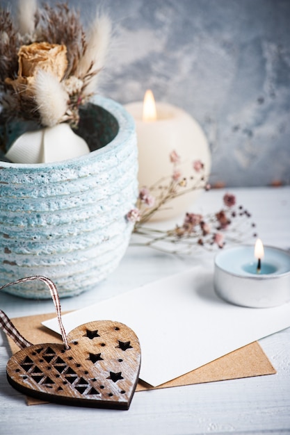 Empty blank note and kraft envelope with heart and lit candles on white wooden table. Copy space for text, for greeting, invitation