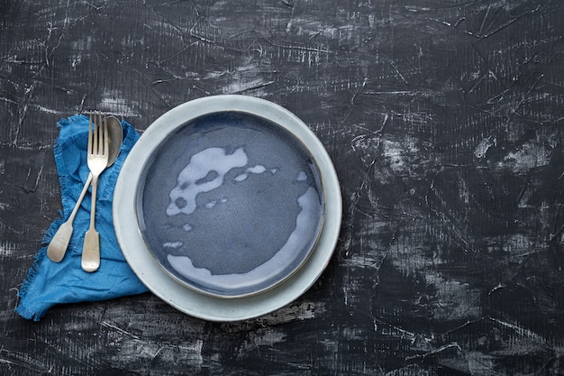 Empty blue plate served with fork spoon table napkin. vintage blue plate for luxury dinner in restaurant. Dark black concrete table chalk board copy space.