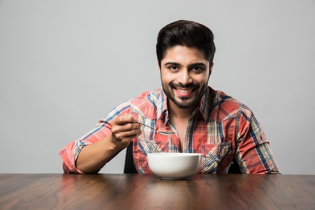 Empty bowl and Indian man with beard holding spoon or chopsticks, wearing checkered shirt and sitting at table