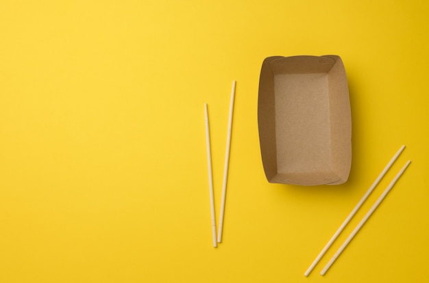 Пустая тарелка коричневой бумаги и деревянные палочки для еды на желтом фоне, вид сверху. Одноразовая посуда, без отходов