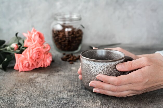 An empty ceramic cup in female hands a jar of coffee beans and a bouquet of roses on the table