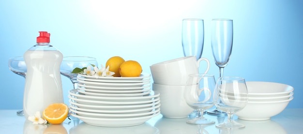 Фото Пустые чистые тарелки, стаканы и чашки с жидкостью для мытья посуды и лимоном на синем фоне