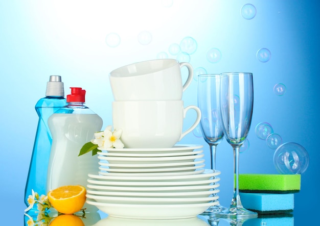 Empty clean plates glasses and cups with dishwashing liquid sponges and lemon on blue background