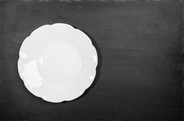 Empty clean white plate with figured wavy edges on black background of shale serving board. Mock up for prepared dish. Copy space for recipe or text. Overhead view. Minimal image.