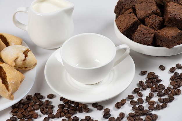 Tazza di caffè vuota in ceramica con brocca di latte torta al cioccolato e chicchi di caffè su sfondo bianco