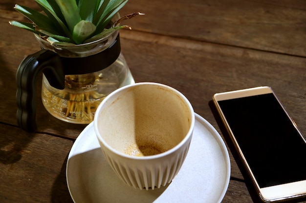 空白の画面のスマートフォンと鉢植えの植物と木製のテーブルでコーヒーを飲み終えた空のカップ