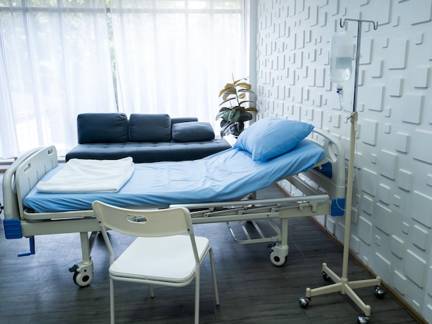 Empty electric adjustable patient bed in a hospital room with blue pillow and bedsheet saline solution bags chair sofa near the window in recovery room Health care medical and insurance concept