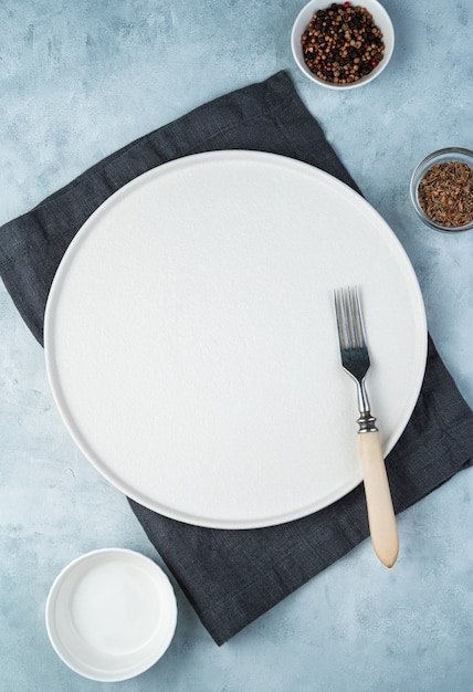 An empty flat white plate, fork, and spices in cups on a gray background.