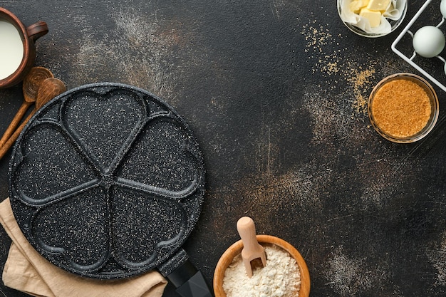 空のフライパンブラック、灰色のコンクリートテーブルの背景に朝食のハートと材料の形をしたパンケーキを焼くための石の焦げ付き防止コーティングが施されたスキレット。バレンタインデーの朝食。上面図。