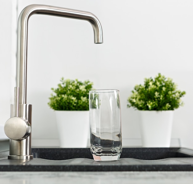 Empty glass in the kitchen sink near the water tap.