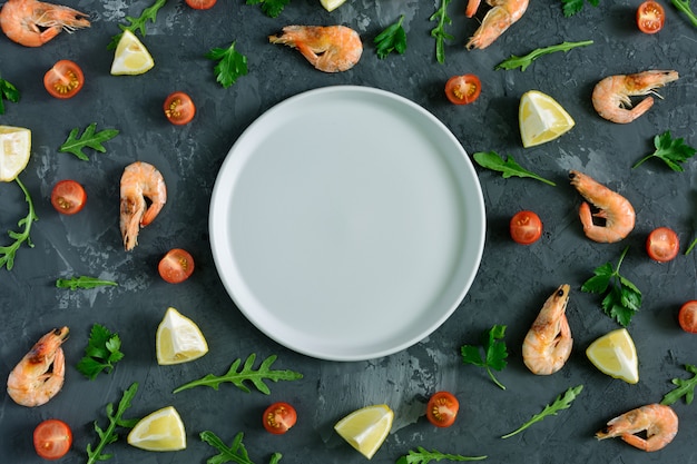 An empty gray plate lies in the center of a dark textured background. Around are scattered lemon, parsley, rucola, cherry tomatoes and shrimp. Photos from the top, mock up.