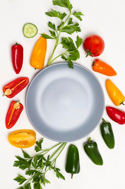 Empty gray plate. Sprigs of parsley, green and red peppers, tomatoes on table. White background. Flat lay.