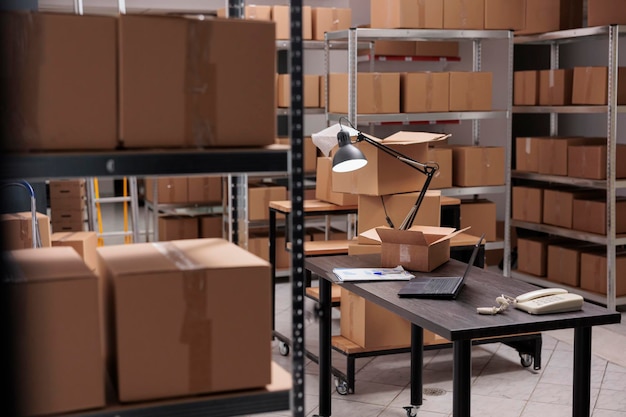 Empty huge distribution storehouse in logistic center with high shelves full with cardboard boxes and desk with shipping documents on it. Concept of heavy industry manufacturing factory.