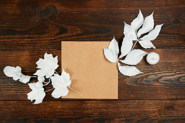 Empty kraft sheet of paper flat lay mockup for your art, picture or hand lettering composition copy space, top view. Autumn composition made of white leaves on dark brown wooden backgound