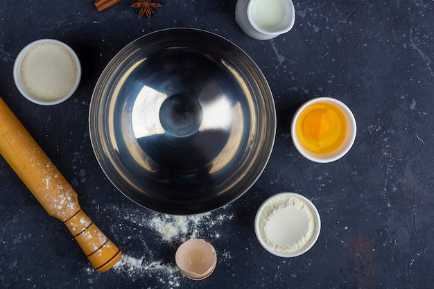 Empty metal bowl among ingredients and utensils for cooking cake (flour, egg, milk, sugar, rolling pin, towel) on dark table. Food concept. Top view, flat lay, copy space for text