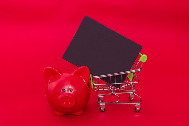 Empty mini chalk board, shopping trolley and piggy bank on red Christmas background. Copy space, mockup