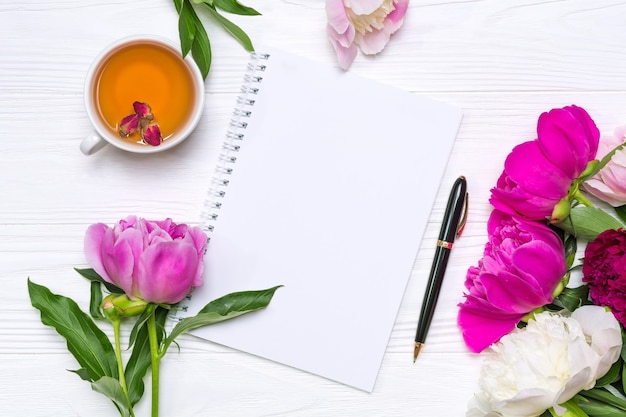 Empty notebook with place for text, pen, a cup of tea and peonies flowers on a white wooden background.