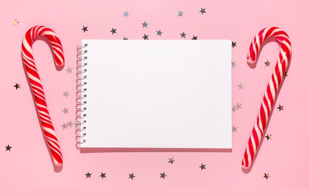 Empty notebook with red Christmas candies and confetti stars pours on a pastel pink background, top view. Flat lay. Holiday new year, greeting or good news concept. Mockup template. Blank notepad