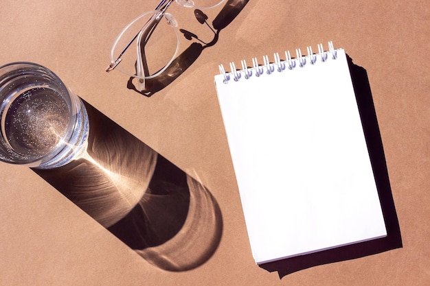 Empty notepad glass of water and eyeglasses with strong shadows on brown background Creative mockup Top view flat lay Still life