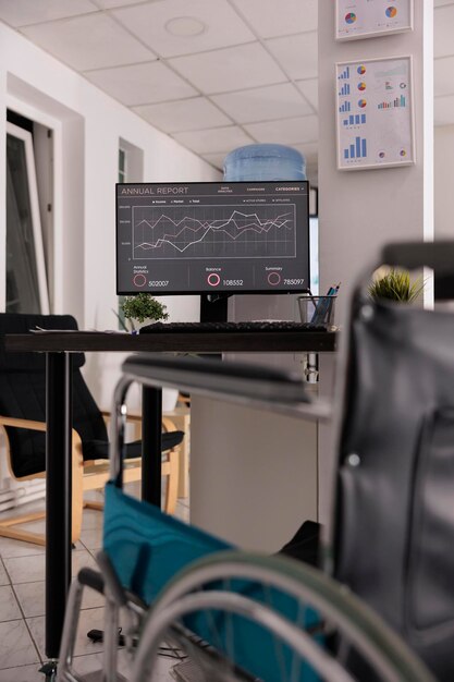 Empty office wheelchair accessible next to desk, company with policies of inclusion and equality. Computer monitor on the table with annual data upload report, interior workplace.