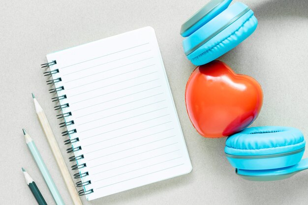 Опорожните открытую страницу тетради белой бумаги с карандашами и красного сердца с наушниками на белой таблице.