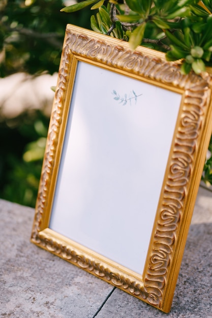 Empty painting in a beautiful carved wooden frame stands on a stone slab against a green olive tree