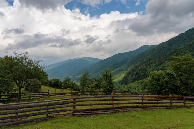 Empty pasture in the mountains fenced with a wooden fence against the backdrop of the forest sky and clouds