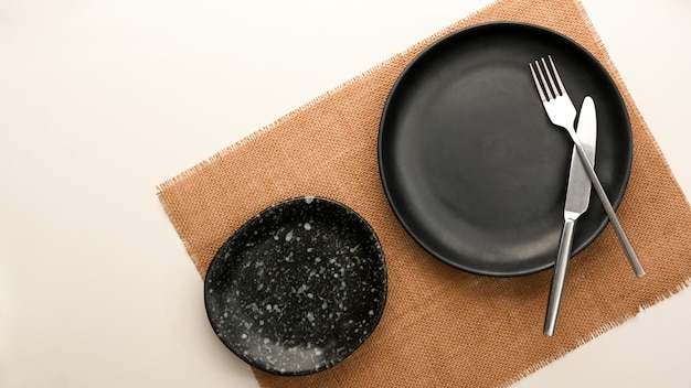 Empty patterned black plate, silverware and brown placemat on white table. flat lay, top view