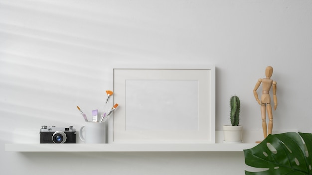 Empty photo frame, camera and decorations on white shelf with white wall