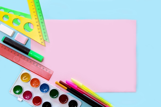 Empty pink sheet on a blue background surrounded by school accessories triangles pens watercolors