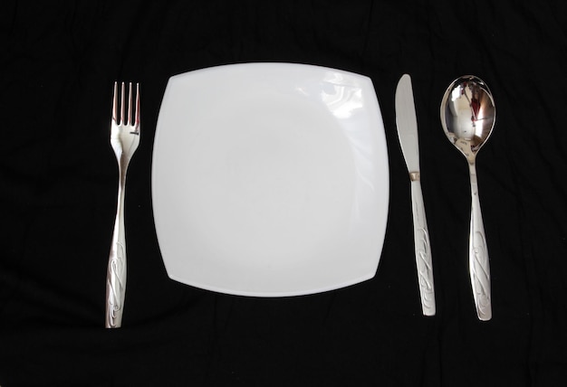 Пустая тарелка, вилка, ложка и нож на черном фоне, чистая тарелка и столовые приборы на черном фоне