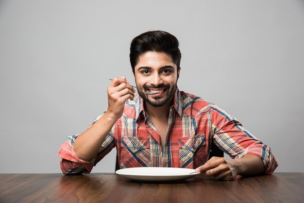 Empty plate and Indian man with beard holding spoon and fork, wearing checkered shirt and sitting at table