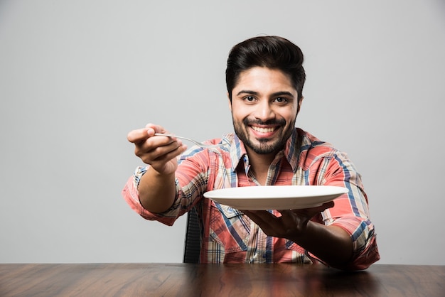 Empty plate and Indian man with beard holding spoon and fork, wearing checkered shirt and sitting at table