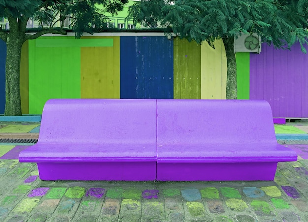 Яркая фиолетовая бетонная скамейка в стиле поп-арт с синим и зеленым старым деревянным зданием в фоновом режиме