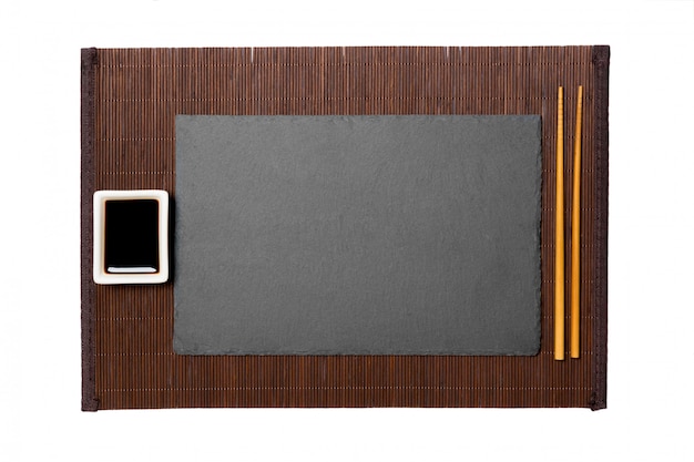 Empty rectangular black slate plate with chopsticks for sushi and soy sauce on dark bamboo mat background.