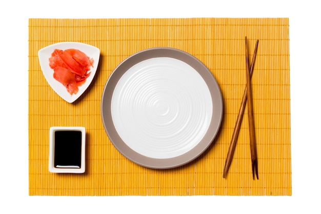 Empty round white plate with chopsticks for sushi, ginger and soy sauce on yellow bamboo mat background. Top view with copy space for you design.