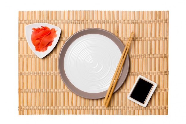 Empty round white plate with chopsticks for sushi and soy sauce, ginger on yellow bamboo mat background. Top view with copy space for you design