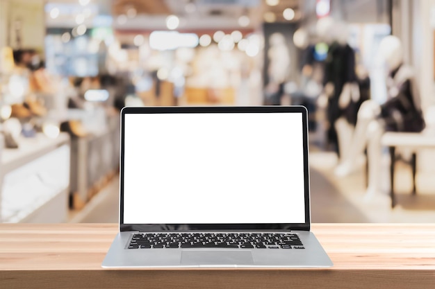 Empty screen laptop on a wooden table with abstract blurred background of interior clothing store at Shopping Mall