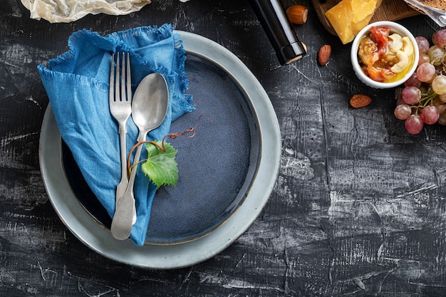 Foto piatto blu da portata vuoto con cucchiaio forchetta in cornice di ingredienti alimentari cucina mediterranea gastronomia antipasto snack uva formaggio vino. piatto piatto blu con copia spazio sul tavolo di cemento scuro.