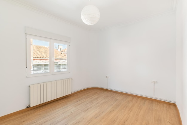 An empty small bright room with white walls and a window overlooking a neighbors house on a sunny