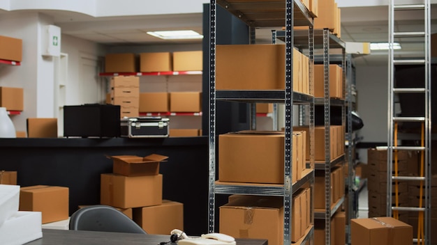 Empty storehouse with office desk and goods to do inventory and ship merchandise to retail clients. Storage room filled with cardboard packages and boxes on storehouse racks, small business.