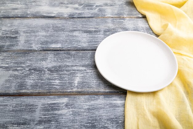 Empty white ceramic plate on gray wooden background and yellow linen textile. Side view,