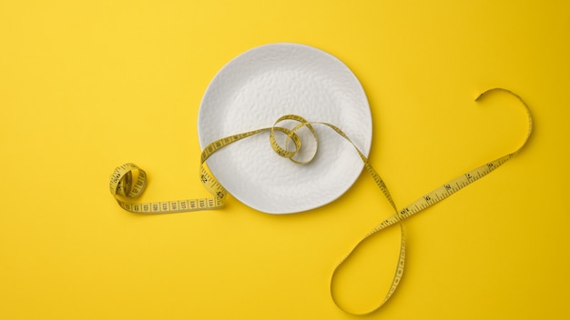 Empty white ceramic plate and measuring tape on yellow background. Healthy nutrition concept, weight loss