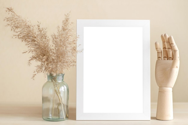 An empty white frame stands on a beige wooden table with dried herbs in a vase and a wooden hand.