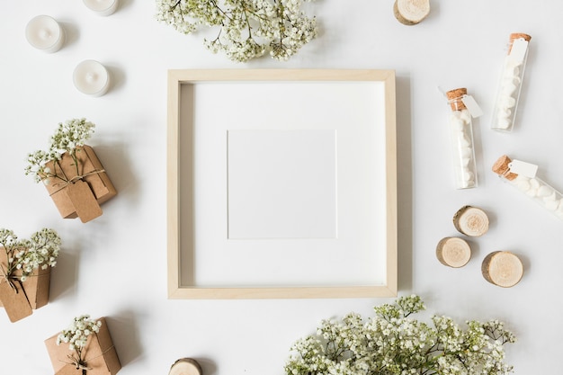 An empty white frame surrounded with gift boxes; candles; tree stump; marshmallow test tubes and baby's-breath flowers on white backdrop