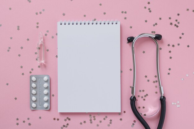 Empty white paper sheet notebook, blister tablet, stethoscope or phonendoscope on a pink with confetti stars