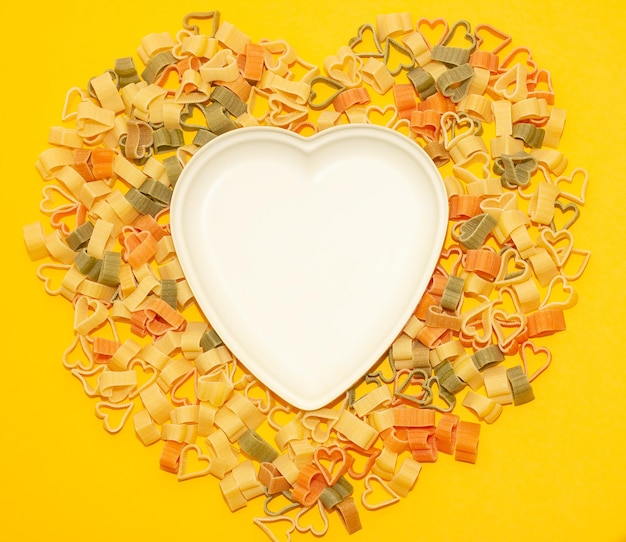 Empty white plate in the shape of a heart on a background of colored pasta