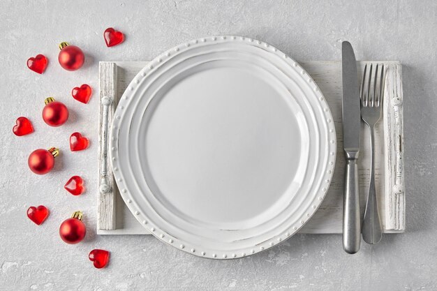 Empty white plate with cutlery on a white wooden tray on a gray concrete table surrounded by red Christmas balls and hearts Template for presenting food on the menu Top view with copy space