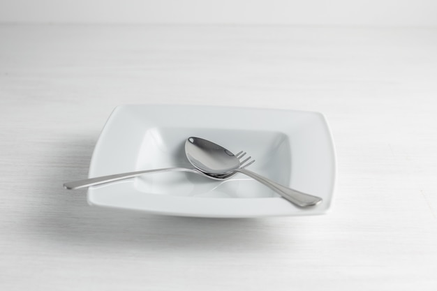An empty white plate with a spoon and fork. Light background