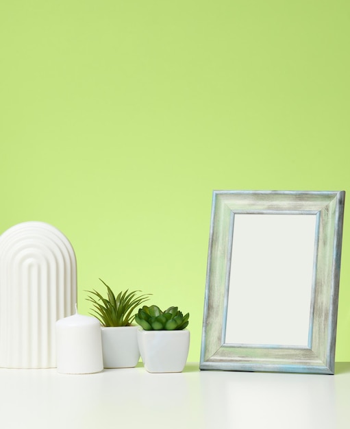 Empty white wooden photo frame and flowerpots with plants on white table, green background